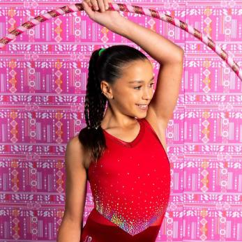 Young girl in the Heart 2 Heart leotard from the Funscape Collection, smiling and posing with a hula hoop against a vibrant pink patterned background.
